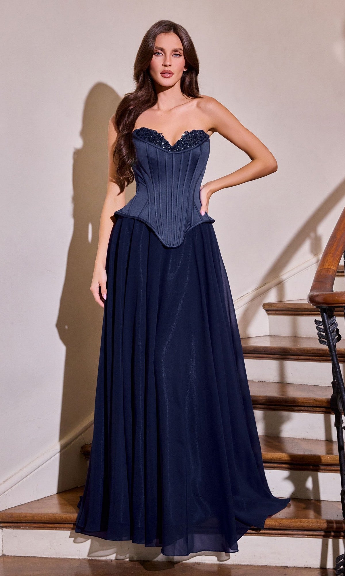 A woman stands indoors beside a stairway, wearing the Ladivine J891 long prom dress in navy. This strapless gown features a corset bodice and flowing chiffon A-line skirt.